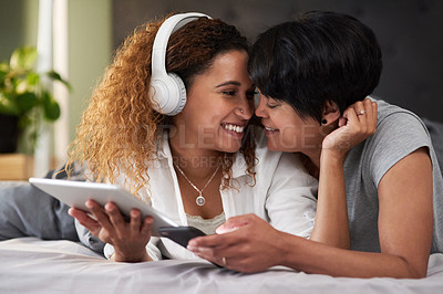 Buy stock photo Shot of a woman wearing headphones and using a digital tablet while lying in bed with her partner