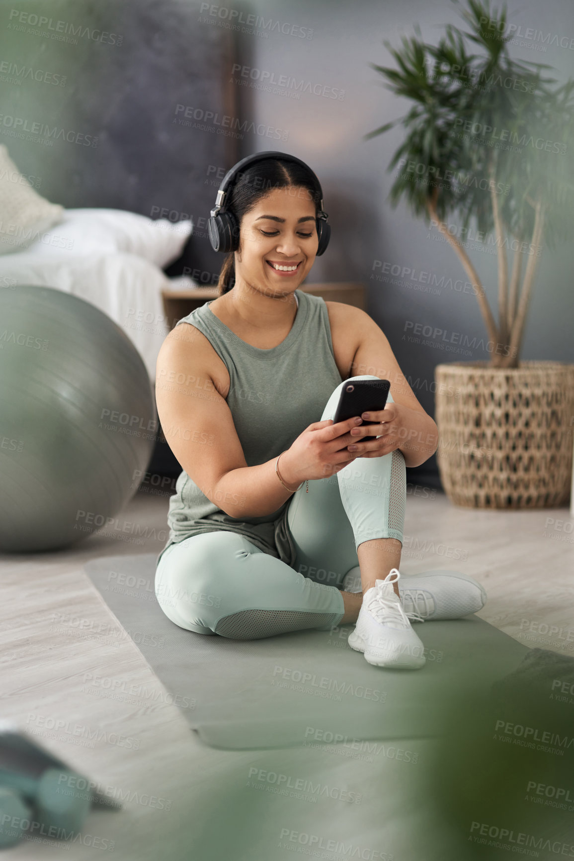 Buy stock photo Shot of a sporty young woman wearing headphones and using a cellphone while exercising at home