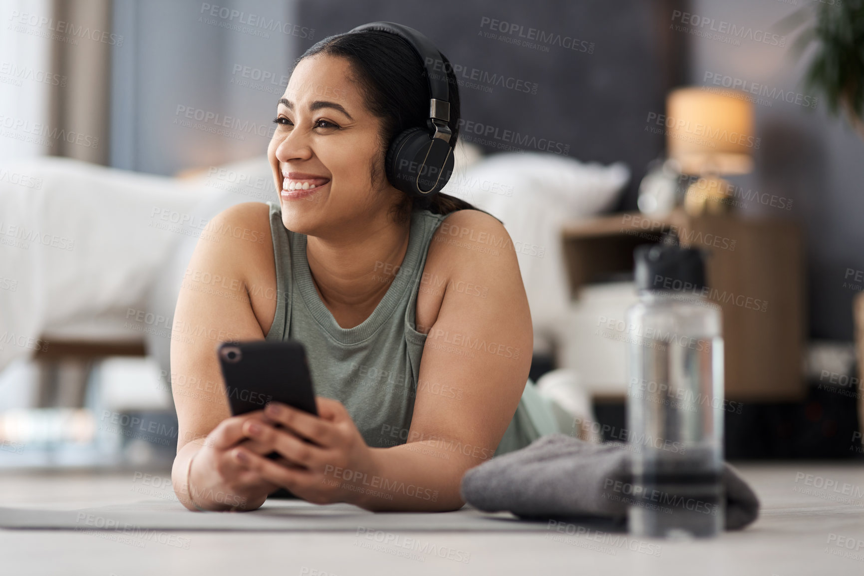 Buy stock photo Shot of a sporty young woman wearing headphones and using a cellphone while exercising at home