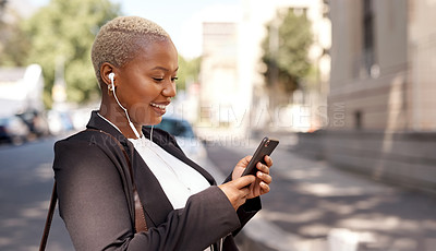 Buy stock photo Shot of a young businesswoman wearing earphones and using a cellphone while out in the city