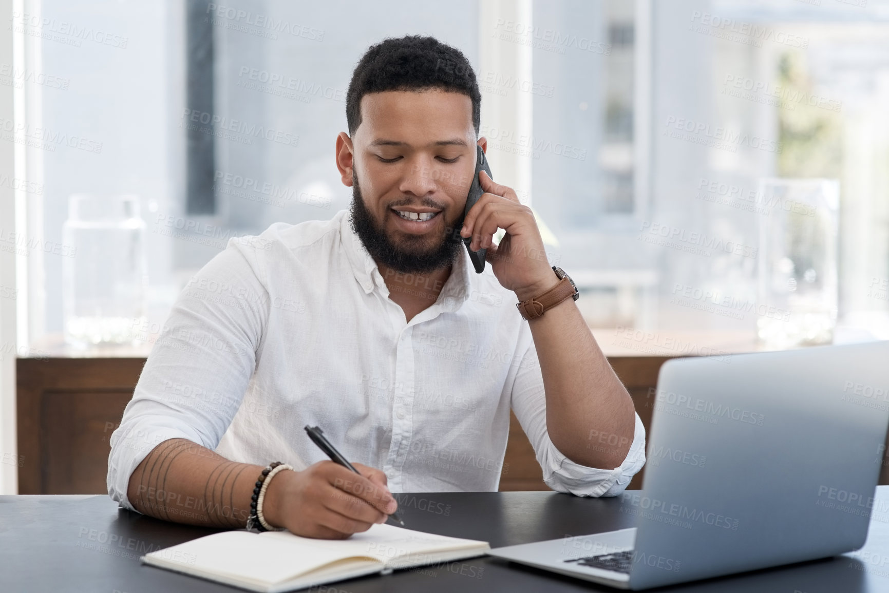 Buy stock photo Shot of a young businessman talking on a cellphone while writing notes and working on a laptop