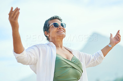 Buy stock photo Shot of an attractive mature woman standing outside alone and raising her hands during a day on the beach