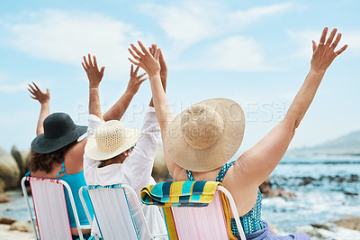 Buy stock photo Shot of an unrecognizable group of friends sitting together and raising her arms during a day on the beach