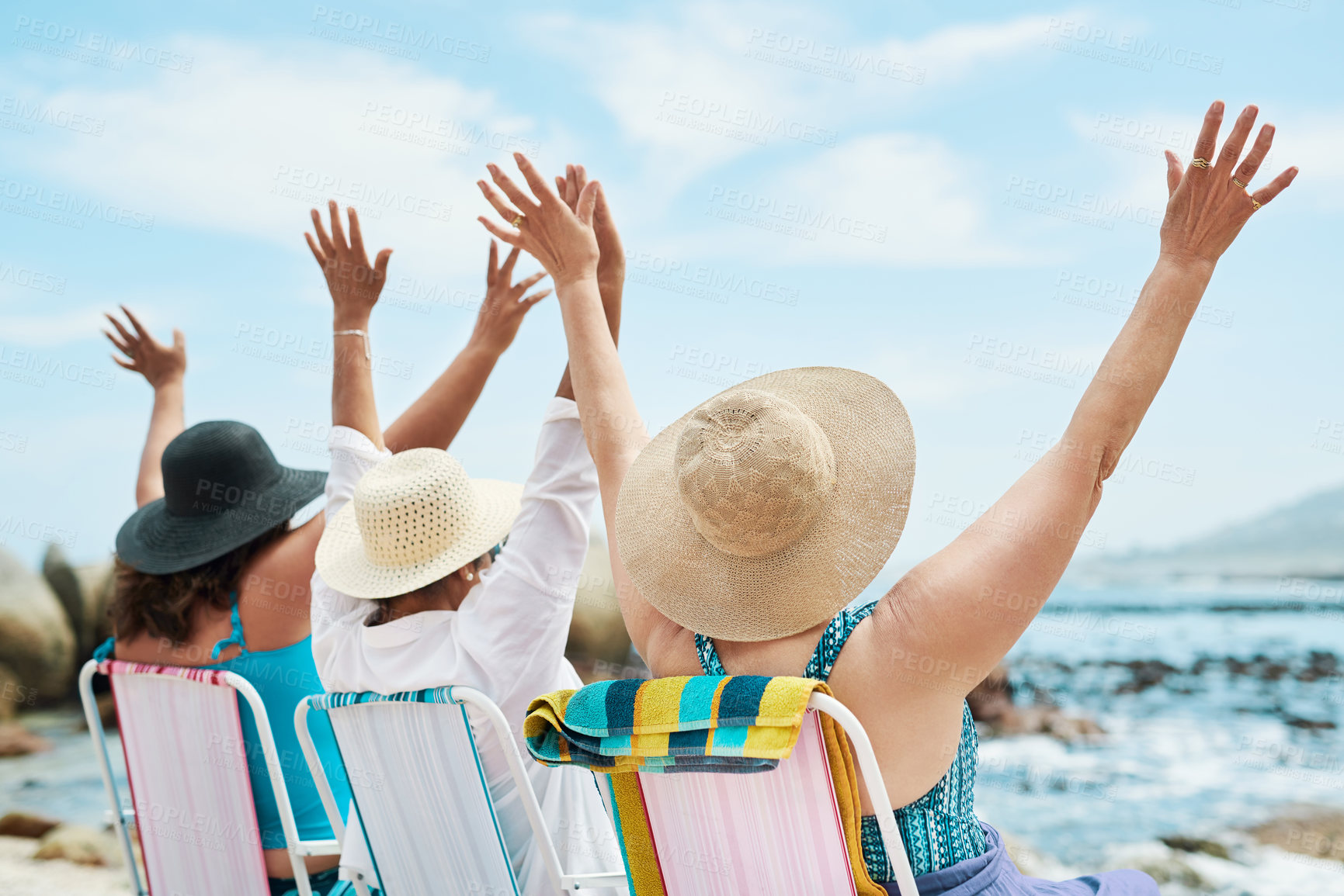 Buy stock photo Shot of an unrecognizable group of friends sitting together and raising her arms during a day on the beach