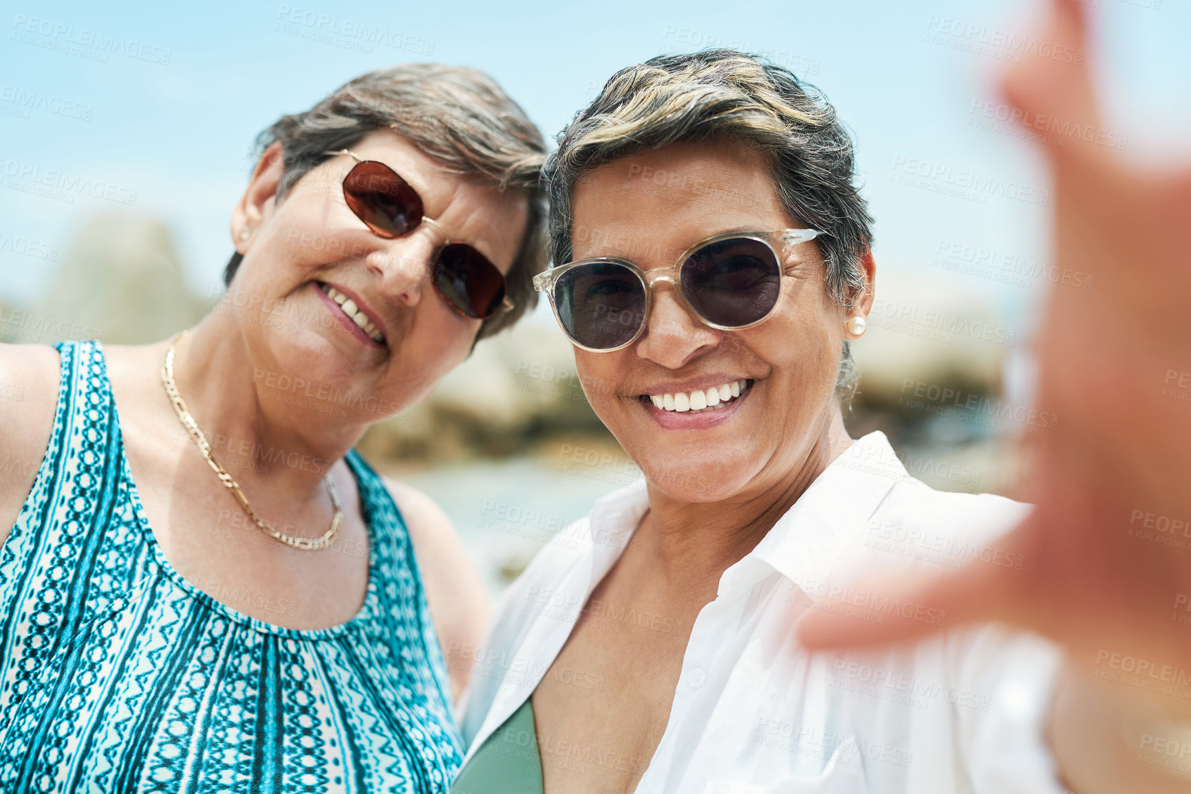 Buy stock photo Shot of two mature friends standing together and posing for a selfie during a day out on the beach