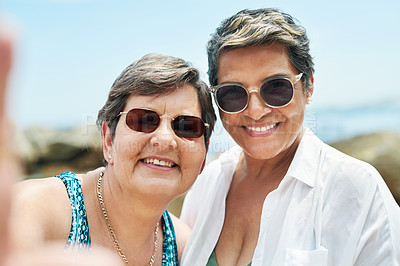 Buy stock photo Shot of two mature friends standing together and posing for a selfie during a day out on the beach