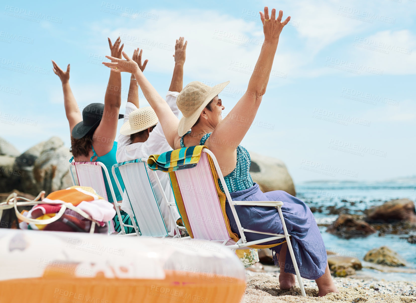 Buy stock photo Shot of a mature group of friends sitting together and raising her arms during a day on the beach