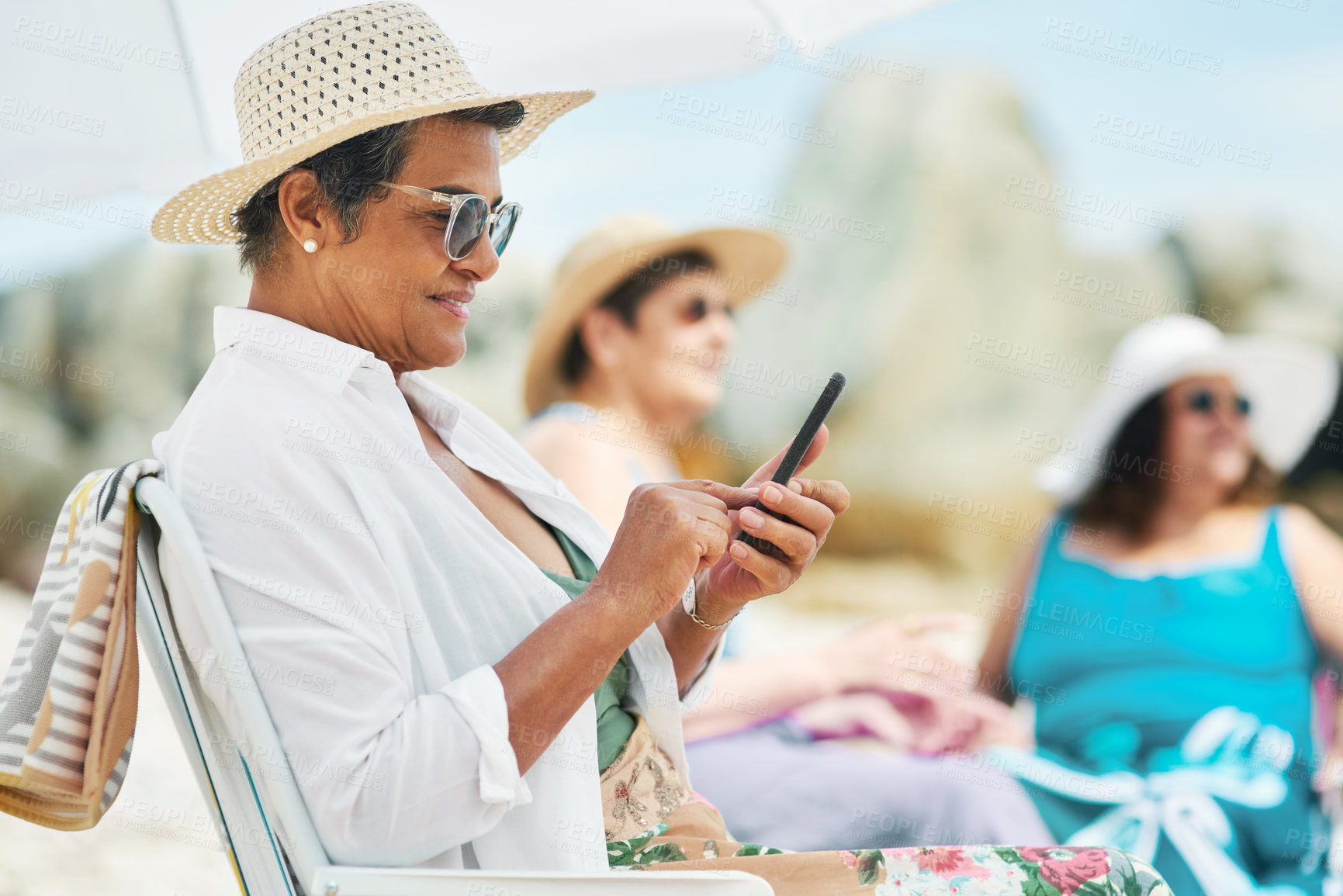 Buy stock photo Shot of a mature woman sitting alone and using her cellphone during a day out on the beach with friends