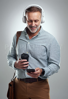 Buy stock photo Studio shot of a mature man wearing headphones and using a cellphone while carrying a bag and cup of coffee against a grey background