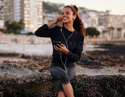 Buy stock photo Shot of an attractive young woman sitting alone on the beach after yoga and listening to music