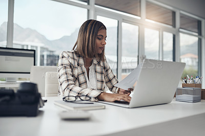 Buy stock photo Shot of an attractive young businesswoman sitting alone in her office and using her laptop while reading paperwork