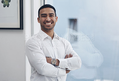 Buy stock photo Shot of a handsome young businessman standing alone in his office with his arms folded during the day