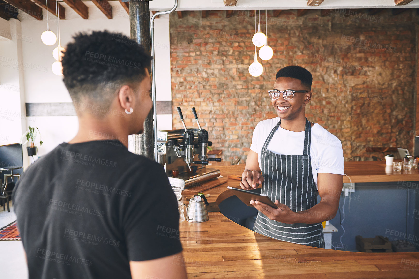 Buy stock photo Shot of a young man using a digital tablet while serving a customer in a cafe