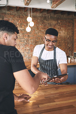 Buy stock photo Shot of a young man using a digital tablet while serving a customer in a cafe
