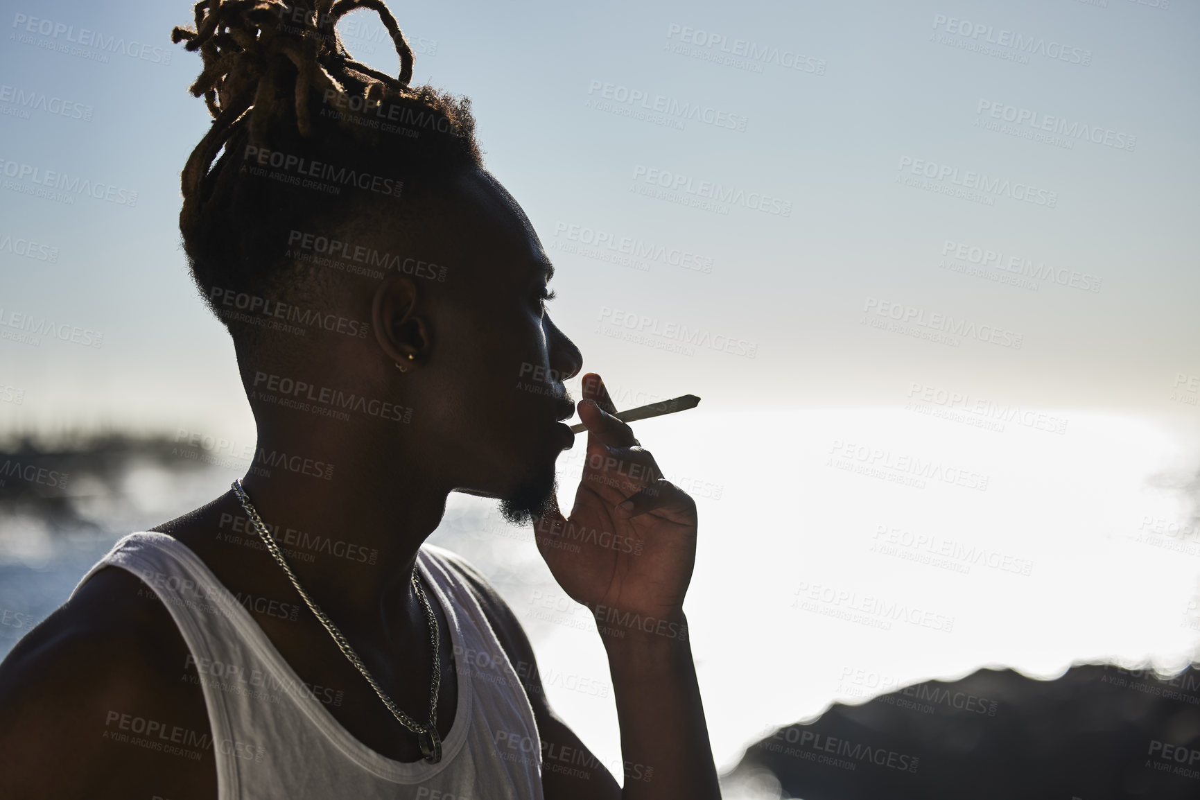 Buy stock photo Shot of a young man smoking a marijuana cigarette at the beach during sunset