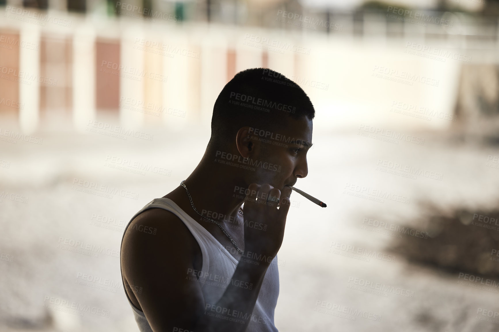 Buy stock photo Shot of a young man smoking a marijuana cigarette against an urban background