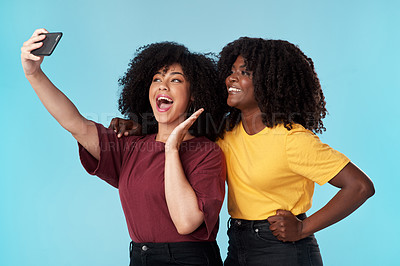 Buy stock photo Studio shot of two young women using a smartphone to take selfies against a blue background