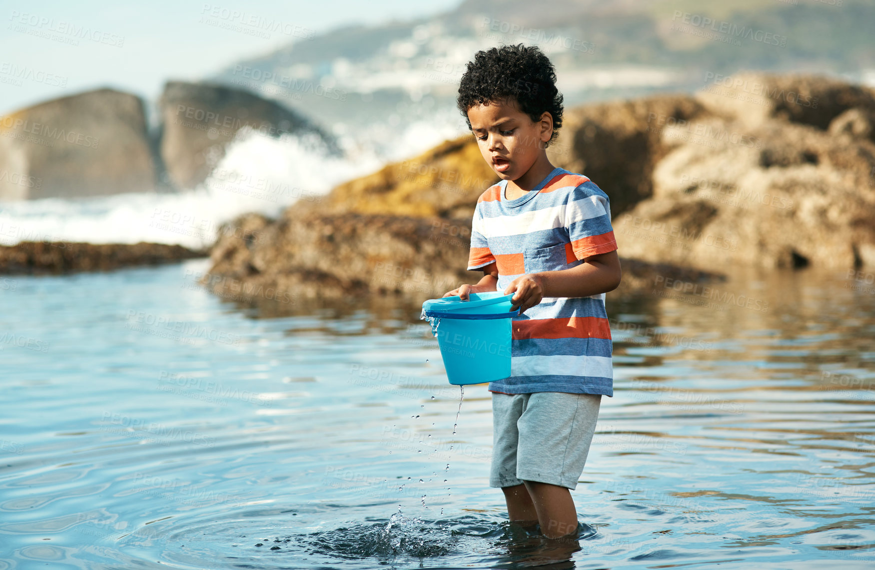 Buy stock photo Shot of a young boy playing in the water at the beach using a bucket to catch things