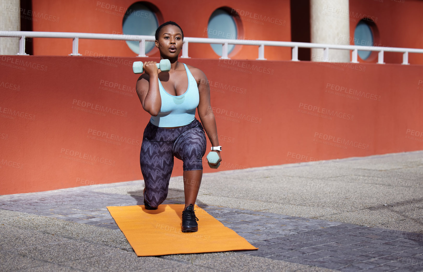 Buy stock photo Shot of a young woman on a gym mat using dummbells against an urban background