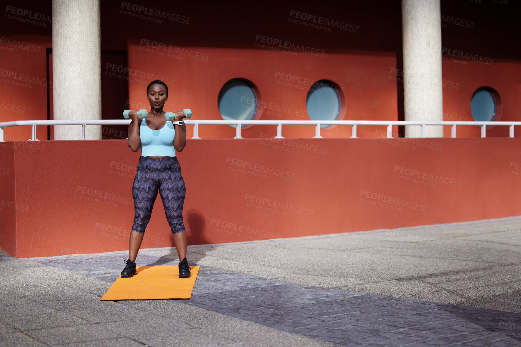 Buy stock photo Shot of a young woman using dummbells on a gym mat against an urban background