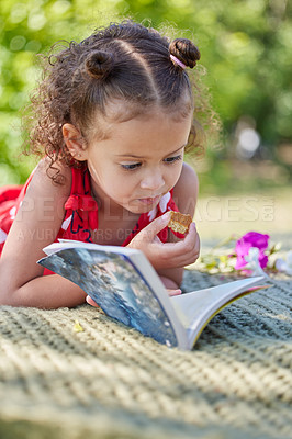 Buy stock photo Girl, child and book on picnic blanket for reading with eating cookies in park to relax with learning. Kid, story and biscuit on lawn for development, education and literacy with sunshine in nature
