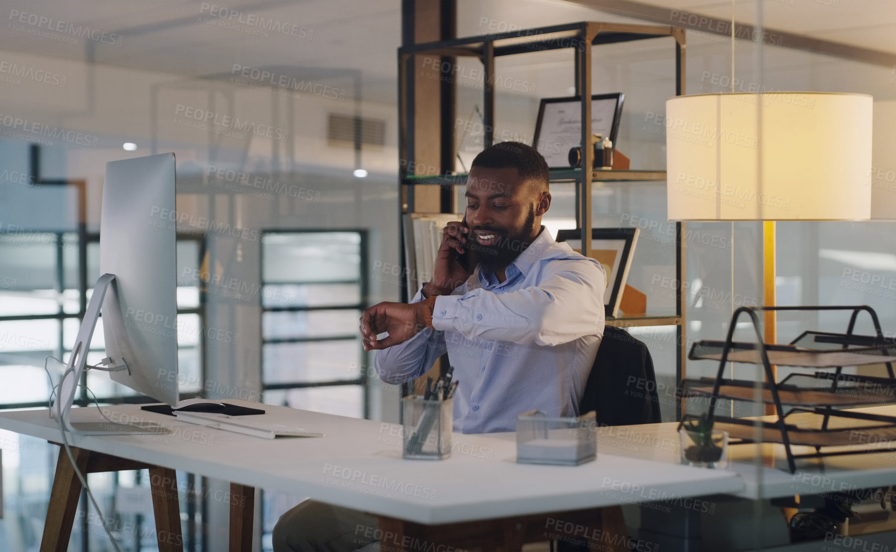 Buy stock photo Shot of a young businessman checking the time on his wristwatch while talking on a cellphone in an office at night