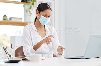Buy stock photo Shot of a young businesswoman wearing a mask and sitting at a desk sanitising her hands in a modern office