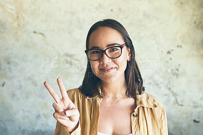 Buy stock photo Portrait of a young woman making a peace sign while standing against a wall
