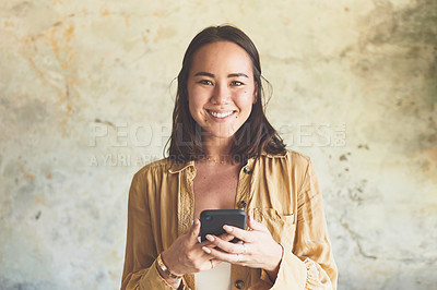 Buy stock photo Portrait of a young woman using a cellphone while standing against a wall