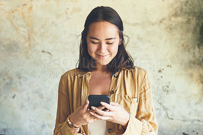Buy stock photo Shot of a young woman using a cellphone while standing against a wall