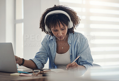 Buy stock photo Shot of an attractive young woman sitting alone in her home and writing in a notebook while wearing headphones