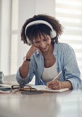 Buy stock photo Shot of an attractive young woman sitting alone in her home and writing in a notebook while wearing headphones