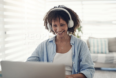 Buy stock photo Shot of an attractive young woman sitting alone at home and using her laptop while wearing headphones
