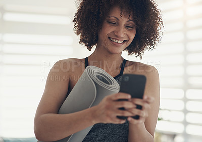 Buy stock photo Shot of an attractive young woman standing in her living room and holding her yoga mat while using her cellphone