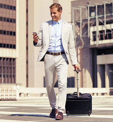 Buy stock photo Shot of a young businessman using a cellphone while walking with a suitcase in the city