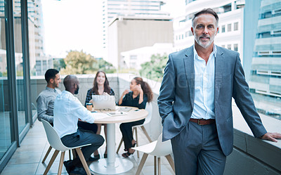 Buy stock photo Portrait of a confident mature businessman standing on the balcony of an office with his colleagues in the background