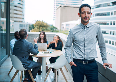 Buy stock photo Portrait of a confident young businessman standing on the balcony of an office with his colleagues in the background