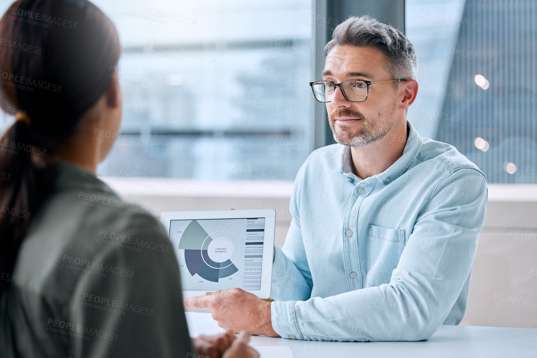 Buy stock photo Shot of a mature businessman speaking to a colleague while analysing graphs on a digital tablet in an office