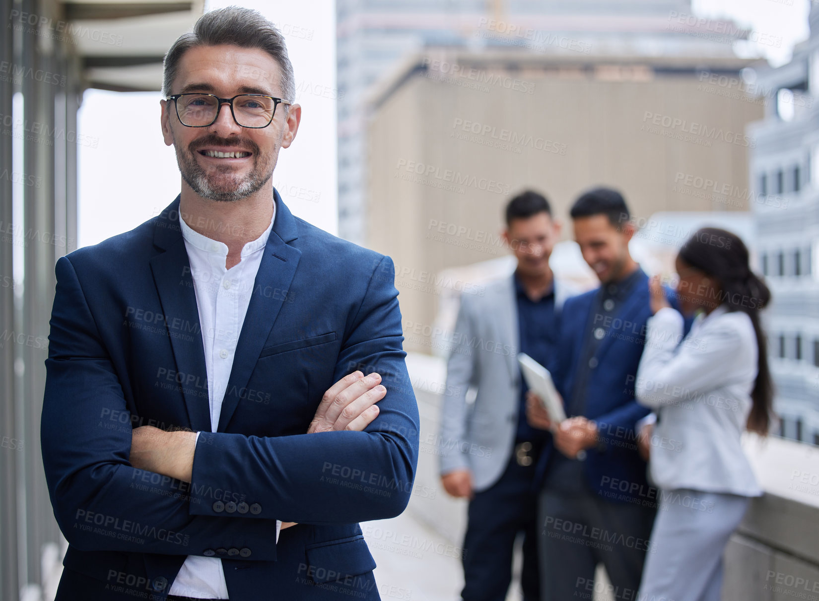 Buy stock photo Cropped portrait of a handsome mature businessman standing outside with his arms folded with his colleagues in the background
