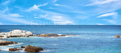 Buy stock photo Boulders at a beach against a cloudy blue sky background with copy space. Calm and majestic ocean across the horizon and rocky coast. Scenic landscape of the sea for a summer holiday or getaway