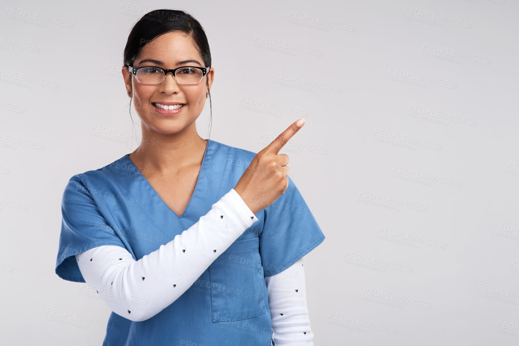 Buy stock photo Portrait of a young doctor wearing glasses and scrubs, pointing to her left against a white background