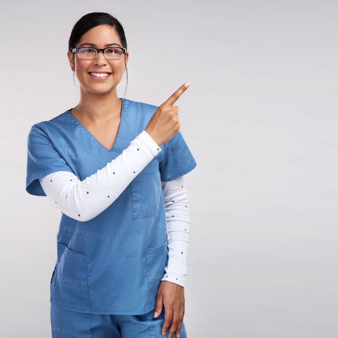 Buy stock photo Portrait of a young doctor wearing glasses and scrubs, pointing to her left against a white background