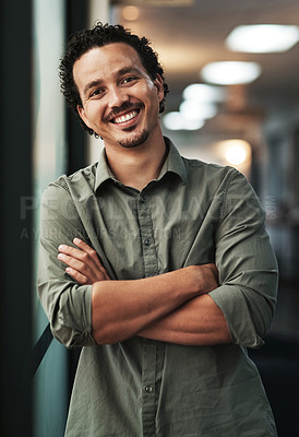 Buy stock photo Shot of a handsome young businessman standing alone in the office with his arms folded
