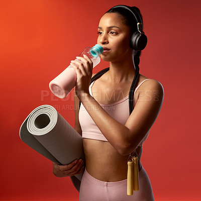 Buy stock photo Studio shot of a sporty young woman drinking water and holding a yoga mat against a red background