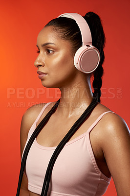 Buy stock photo Studio shot of a sporty young woman wearing headphones and posing with a skipping rope around her neck against a red background