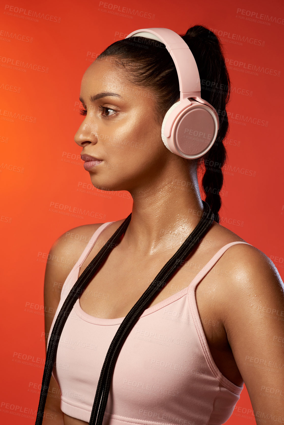 Buy stock photo Studio shot of a sporty young woman wearing headphones and posing with a skipping rope around her neck against a red background