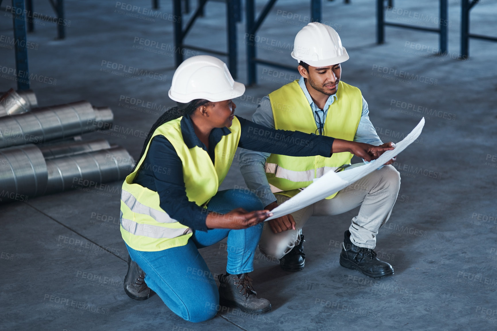 Buy stock photo Shot of two young contractors crouching down in the warehouse together and using a blueprint to plan