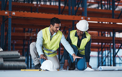 Buy stock photo Shot of two young contractors crouching down in the warehouse together and using a blueprint to plan