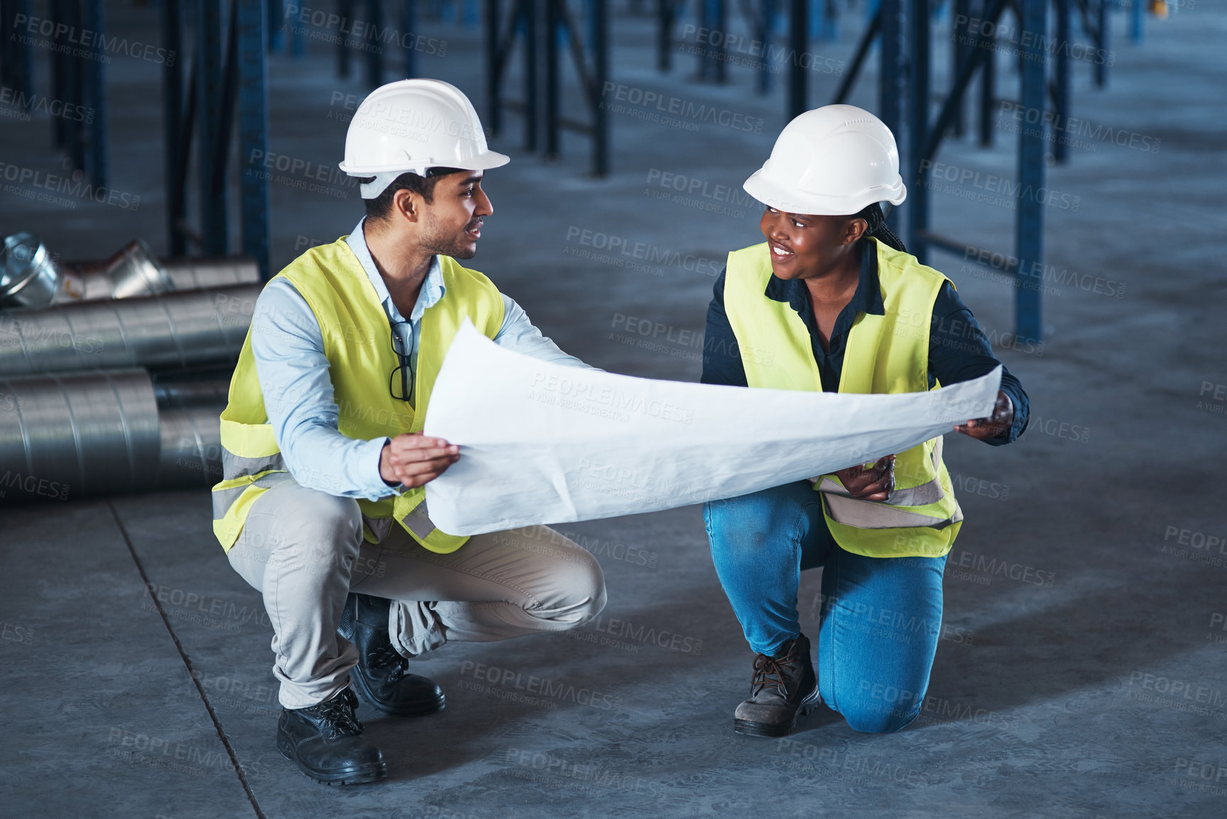 Buy stock photo Shot of two young contractors crouching down in the warehouse together and using a blueprint to plan