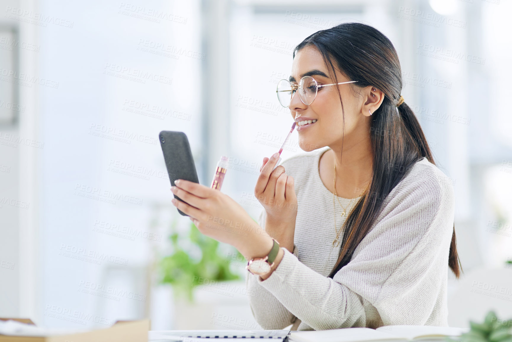 Buy stock photo Shot of a young businesswoman applying lipstick while using her cellphone as a mirror in an office